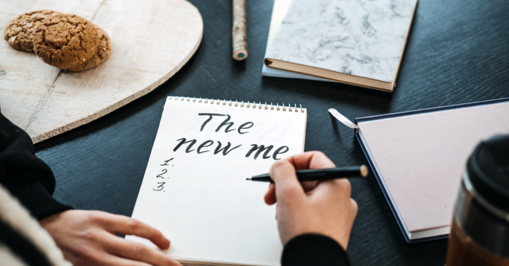 A hand writing "The new me" on a notebook, creating a list. This represents a moment of self-reflection and goal setting, as the individual contemplates personal transformation.