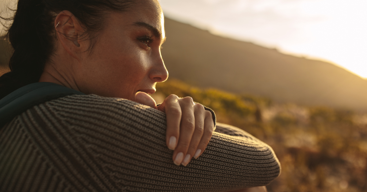 A close-up of a woman in an outdoor setting, deep in thought while gazing into the distance. The soft sunlight and relaxed posture evoke a sense of calm and self-reflection.