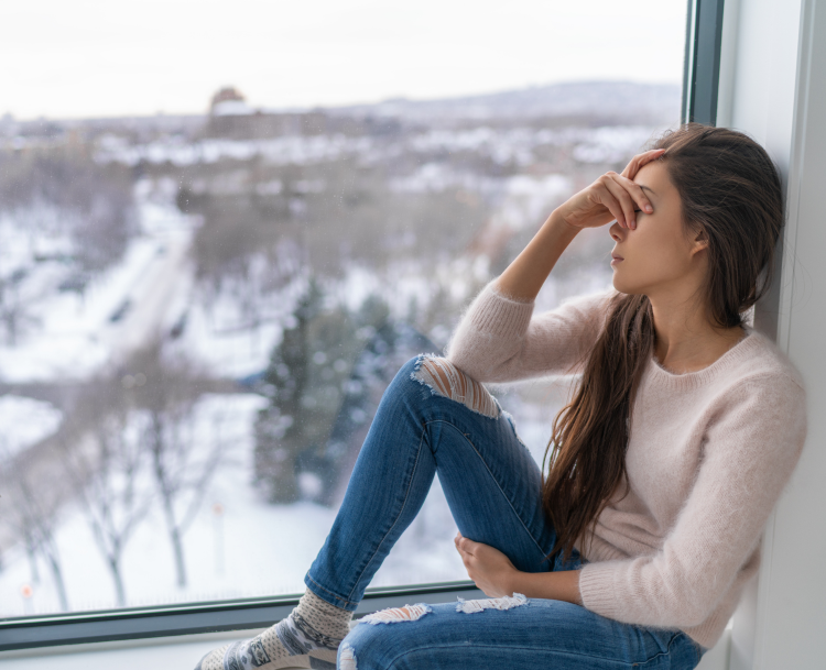 Young woman sitting by a window on a winter day, looking distressed and thoughtful, with a snowy landscape outside.