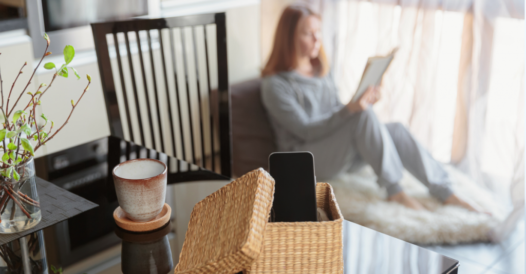 Teen enjoying a digital detox, relaxing with a book on a cozy chair, while a phone rests in a woven basket on a nearby table.