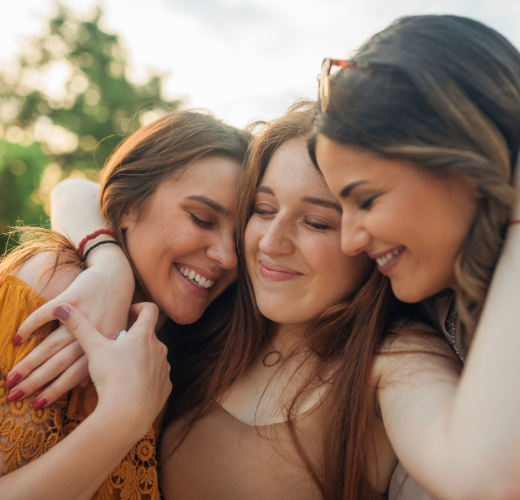 Three women embracing outdoors with closed eyes and gentle smiles, representing healing from PTSD through connection and emotional closeness.