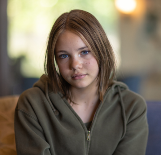 A teen with a calm expression sits in a warmly lit room, wearing a green hoodie. This image reflects the tone of the blog Helping Families Practice Trauma-Informed Care At Home.